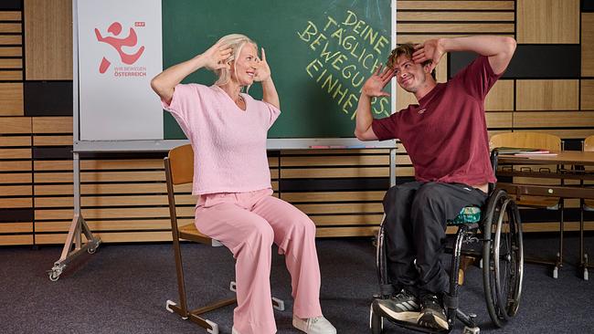 Das Bild zeigt zwei Personen, die in einem Raum mit einer Tafel im Hintergrund sitzen. Die Person ist links sitzt auf einem Stuhl und trägt ein rosafarbenes Outfit, bestehend aus einem Oberteil und einer weiten Hose. Die Person rechts sitzt in einem Rollstuhl und trägt ein weinrotes T-Shirt und dunkle Hose. Bei der linken Person handelt es sich um Karin Augeneder, eine diplomierte Sportlehrerin, bei der rechten Person um Nico Langmann, Profi-Tennisspieler. Beide Personen führen eine Übung aus, bei der sie ihre Hände an die Seiten ihres Kopfes legen. Auf der Tafel im Hintergrund steht in gelber Schrift: „DEINE TÄGLICHE BEWEGUNGSEINHEIT“. Karin und Nico lächeln sich dabei an. Links neben der Tafel ist ein Plakat mit dem Logo „ORF WIR BEWEGEN ÖSTERREICH“ zu sehen. Rechts steht ein Schreibtisch mit zwei Sesseln. Der Raum hat eine moderne Holzwandverkleidung und einen blauen Teppichboden.