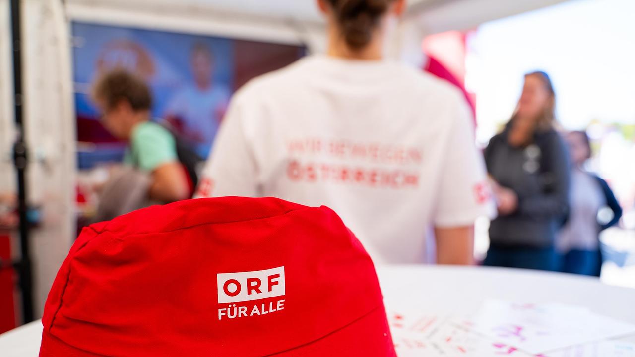 Eine roter Fischerhut mit der Aufschrift „ORF für alle“ liegt im Vordergrund auf einem Tisch. Im Hintergrund ist eine Person zu sehen, die ein „Wir bewegen Österreich“-T-Shirt trägt.