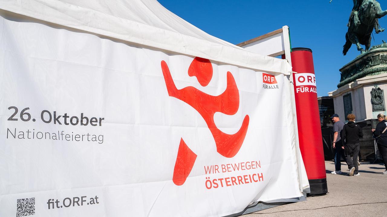 Ein großes Banner mit dem Logo von „Wir bewegen Österreich“ und der Aufschrift „26. Oktober – Nationalfeiertag“ ist an einem Zelt befestigt. Im Hintergrund steht eine Statue auf einem Platz.