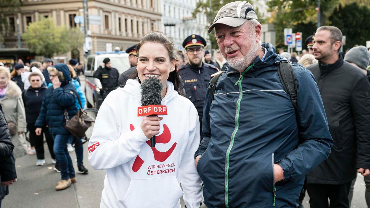 © ORF/Thomas Jantzen Conny Kreuter mit einem Teilnehmer der Ringmeile. Conny trägt einen weißen Hoodie mit rotem WIR BEWEGEN ÖSTERREICH Logo und hält ein ORF-Mikrofon.