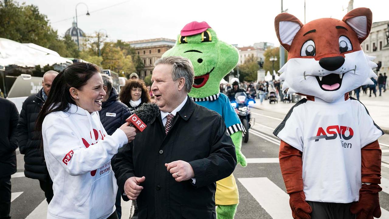 © ORF/Thomas Jantzen Conny Kreuter interviewt Bürgermeister Michael Ludwig während der "Ringmeile". Im Hintergrund sind die Maskottchen von Sportvereinen zu sehen.