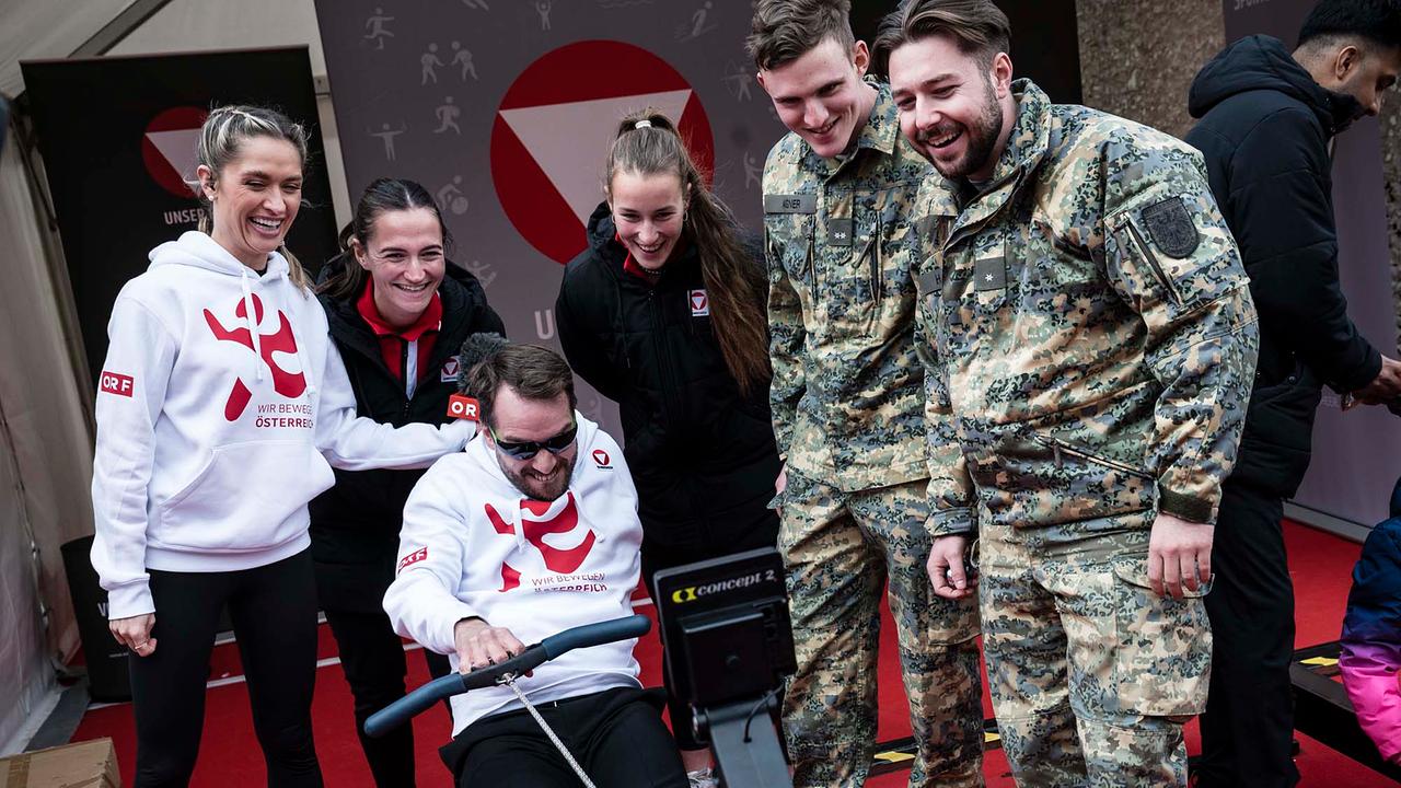 © ORF/Thomas Jantzen Andreas Onea auf einem Rudergerät, neben ihm stehen Corinna Kamper und zwei Soldaten des Bundesheeres. Corinna und Andreas tragen weiße Hoodies mit dem roten WIR BEWEGEN ÖSTERREICH Logo.