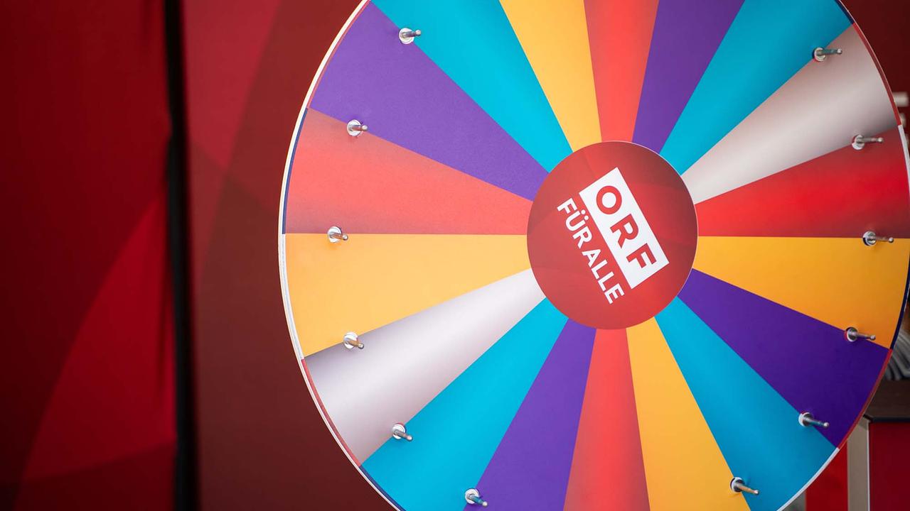 © ORF/Osman Cetin Das Bild zeigt ein buntes Glücksrad mit dem Schriftzug „ORF FÜR ALLE“ in der Mitte. Das Rad ist in verschiedene farbige Segmente unterteilt, unter anderem in Gelb, Orange, Rot, Violett, Blau und Türkis. Am Rand sind kleine Metallstifte befestigt, die als Stopper für das Drehen dienen. Der Hintergrund ist in Rottönen gestaltet.