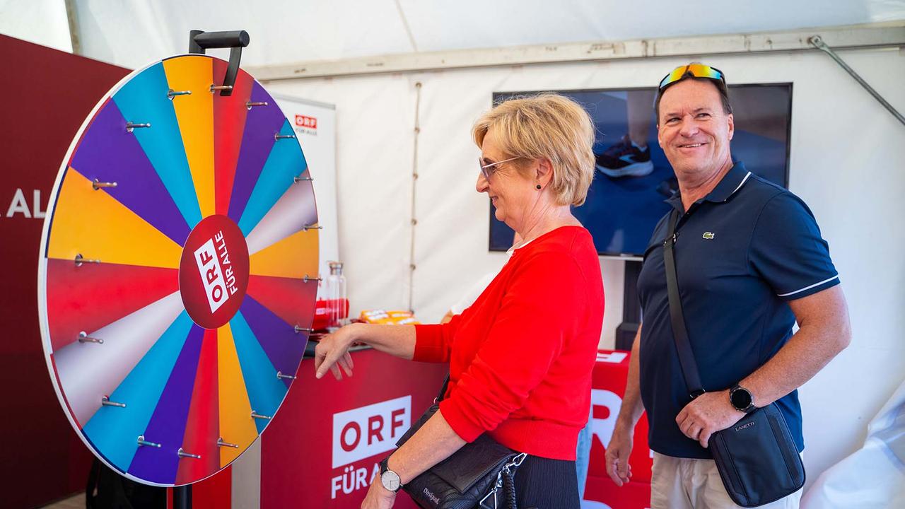 © ORF/Osman Cetin Das Bild zeigt eine Dame in einem roten Pullover, die an einem Glücksrad dreht. Das Rad ist in verschiedene farbige Segmente unterteilt, unter anderem in Gelb, Orange, Rot, Violett, Blau und Türkis. Am Rand sind kleine Metallstifte befestigt, die als Stopper für das Drehen dienen. Der Hintergrund ist in weißen und roten Tönen gestaltet. Neben ihr steht ein Mann.
