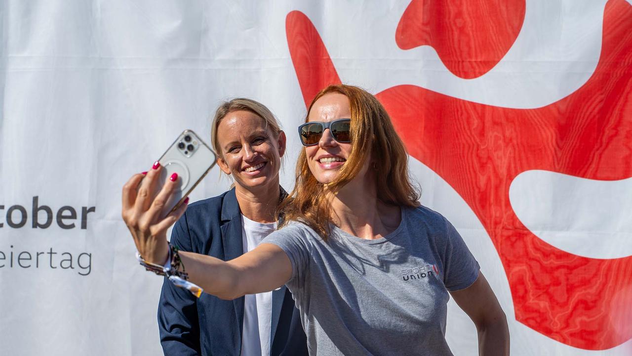 © ORF/Osman Cetin Eine Person hält ein helles Smartphone mit drei Kameralinsen hoch und macht ein Selfie. Hinter ihr steht eine weitere Person in einem dunkelblauen Blazer über einem weißen Oberteil vor einem "WIR BEWEGEN ÖSTERREICH"-Banner. Die Szene spielt im hellen Sonnenlicht.