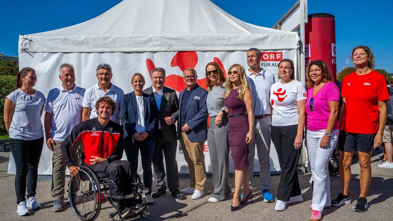 © ORF/Osman Cetin Eine größere Gruppe posiert bei strahlendem Sonnenschein vor einem weißen Pavillonzelt mit ORF‑Branding WIR BEWEGEN ÖSTERREICH und dem Schriftzug „ORF FÜR ALLE“. Im Vordergrund links sitzt Nico Langmann in einem schwarzen Sportrollstuhl und trägt eine rot‑schwarze Trainingsjacke. Dahinter stehen eng beisammen mehrere Menschen in gemischter Kleidung: weiße und rote Sportshirt, Freizeitoutfits, Blazer und Anzüge; eine Frau trägt ein dunkelviolettes Kleid und hohe Schuhe. Rechts ragen zwei rote, zylindrische ORF‑Säulen ins Bild.