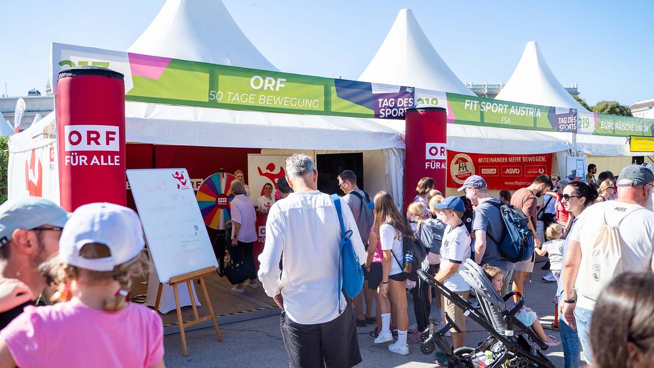 © ORF/Osman Cetin Vor einer Reihe weißer Pagodenzelte mit grünen Bannern „ORF – 50 Tage Bewegung“, „Tag des Sports“ und „Fit Sport Austria“ drängt sich eine große Menschenmenge bei strahlendem Sonnenschein. Am ORF‑Stand markieren zwei rote, zylindrische Säulen mit „ORF FÜR ALLE“ den Eingang; neben der Öffnung steht ein aufgestelltes Whiteboard mit Programmhinweisen. Im Inneren sind ein buntes Glücksrad und Aktionsmaterial zu sehen, Mitarbeitende betreuen die Station. Davor bilden Besucherinnen und Besucher eine lose Schlange, viele mit Rucksäcken und Kappen, Familien mit Kinderwagen und Kindern, die neugierig ins Zelt schauen. Rechts hängen zusätzliche Infobanner, darunter „Miteinander mehr bewegen“ von Verbänden.
