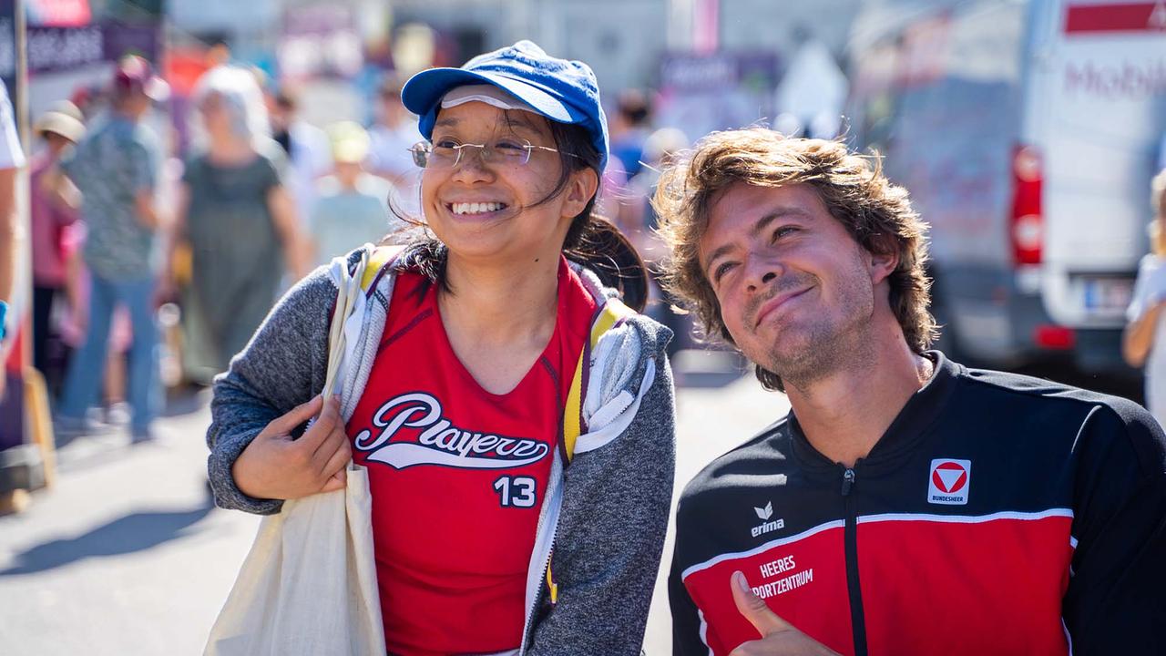 © ORF/Osman Cetin Zwei Personen posieren bei strahlendem Sonnenschein eng beieinander auf einer belebten Fläche am Tag des Sports. Links trägt die Person eine blaue Kappe und ein rotes Sport-Shirt, rechts ist Nico Langmann zu sehen. Beide lächeln für ein Foto.