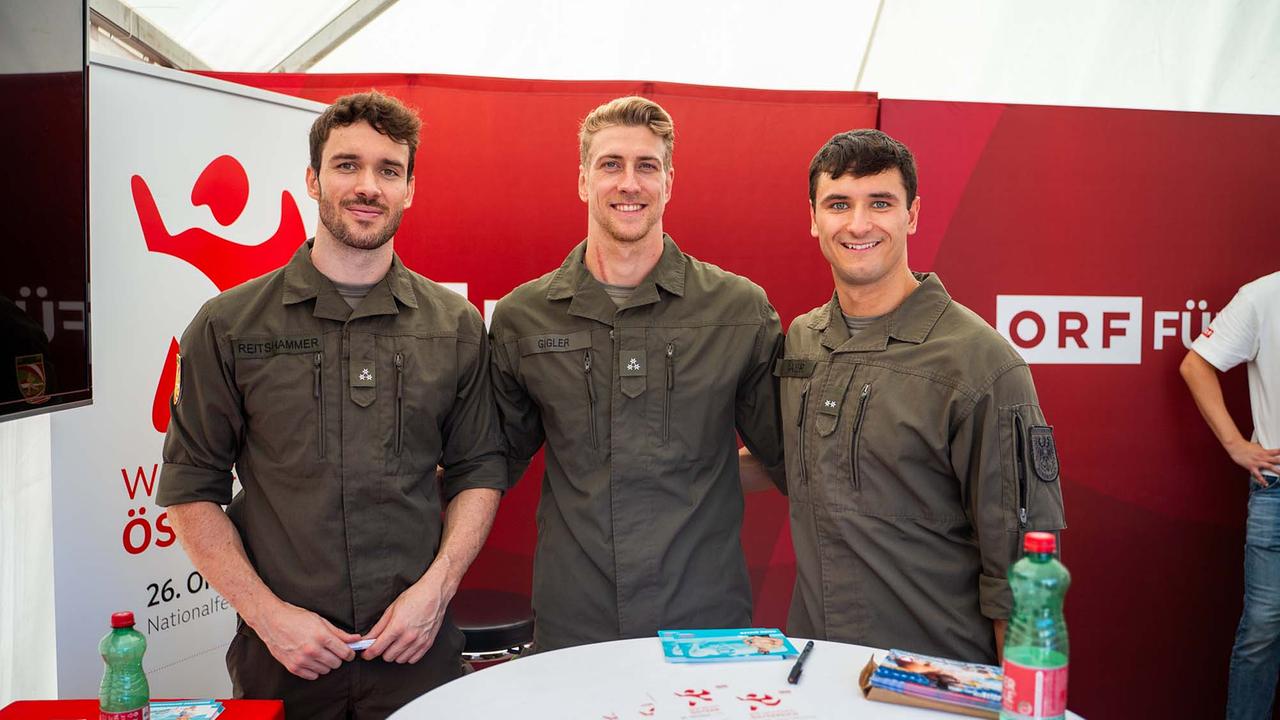 © ORF/Osman Cetin Drei Personen in grüner Bundesheer-Uniformen stehen Schulter an Schulter in einem ORF Eventzelt vor einer rot gestalteten Rückwand mit „ORF FÜR ALLE“. Die Uniformen tragen Namensstreifen, Abzeichen und Taschen auf der Brust, an den Ärmeln sind weitere Embleme zu sehen