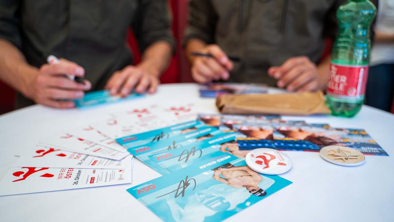 © ORF/Osman Cetin Auf einem weißen runden Stehtisch liegen Infomaterialien und Autogrammkarten, während zwei Personen in grüner Bundesheer-Uniform mit schwarzen Stiften unterschreiben. Sichtbar sind weiße Flyer mit rotem Bewegungslogo und dem Hinweis auf den 26. Oktober/Nationalfeiertag, daneben türkisfarbene Karten eines Schwimmathleten mit bereits gesetzten Signaturen.