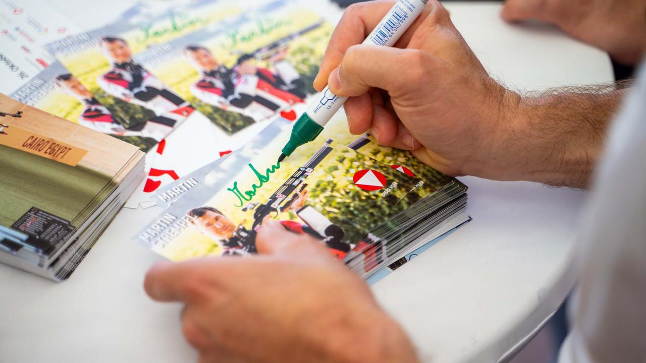 © ORF/Osman Cetin Auf einem weißen Stehtisch werden Autogrammkarten mit einem grünen Filzstift signiert. Die Karten zeigen einen Sportschützen mit Gewehr vor einer sommerlichen Landschaft sowie rot‑weiße Embleme; ein ganzer Stapel liegt bereit, mehrere Exemplare tragen bereits die grüne Unterschrift.