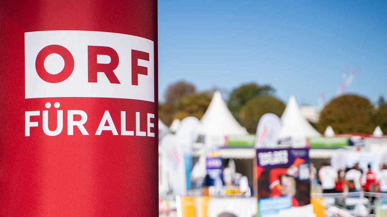 © ORF/Osman Cetin Das Bild zeigt im Vordergrund eine rote Säule mit dem weißen Schriftzug „ORF FÜR ALLE“. Im Hintergrund, leicht unscharf, sind mehrere weiße Zelte, Infostände und Besucherinnen und Besucher eines Outdoor-Events zu sehen. Der Himmel ist blau, und im Hintergrund erkennt man Bäume.