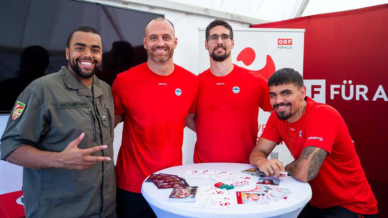 © ORF/Osman Cetin In einem hellen ORF‑Eventzelt stehen vier Personen dicht beisammen hinter einem weißen Stehtisch. Links trägt jemand eine grüne Bundesheer Uniform mit Ärmelabzeichen, daneben posieren drei Personen in roten Sportshirts.