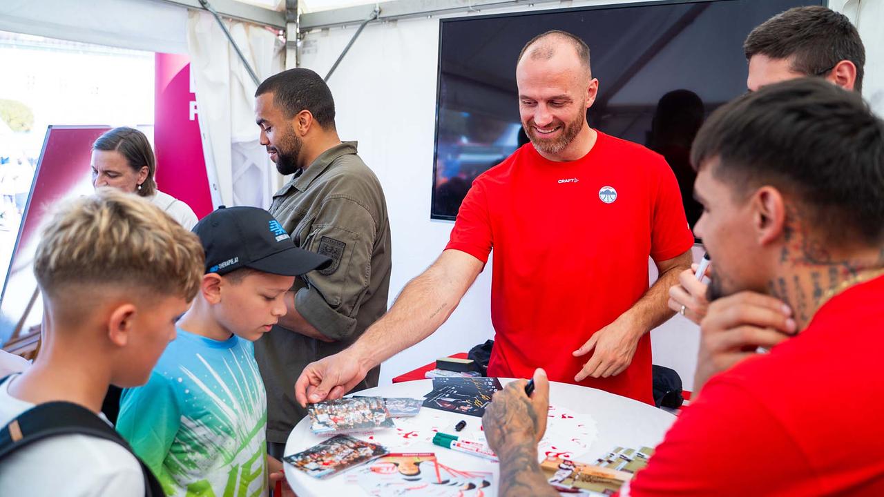 © ORF/Osman Cetin In einem hellen Eventzelt stehen mehrere Menschen dicht um einen runden weißen Stehtisch. Eine Person im roten Sportshirt reicht wartenden Kindern signierte Fotos hinüber, eine weitere mit tätowiertem Arm hält einen Stift bereit.
