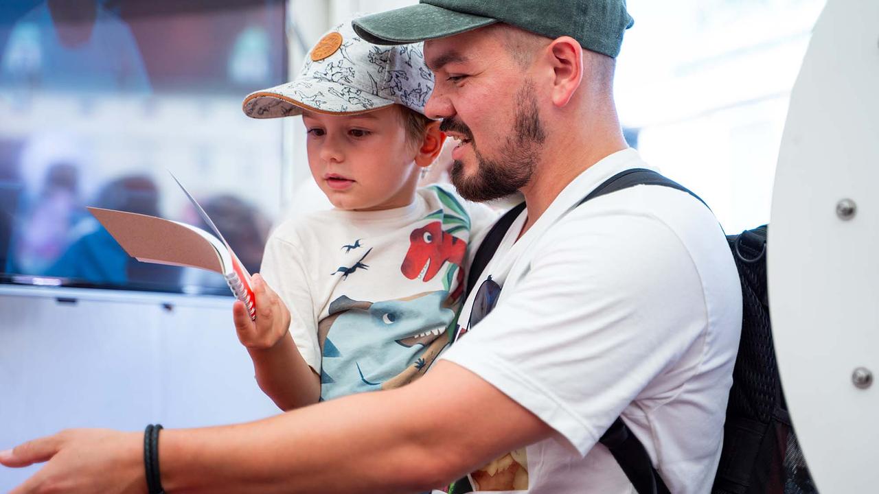 © ORF/Osman Cetin In einem hellen Eventzelt hält eine erwachsene Person mit grüner Kappe und weißem T Shirt ein Kind auf dem Arm. Das Kind trägt ein helles Shirt mit Dinosaurier Motiven und hält einen kleinen Block und einen Stift.