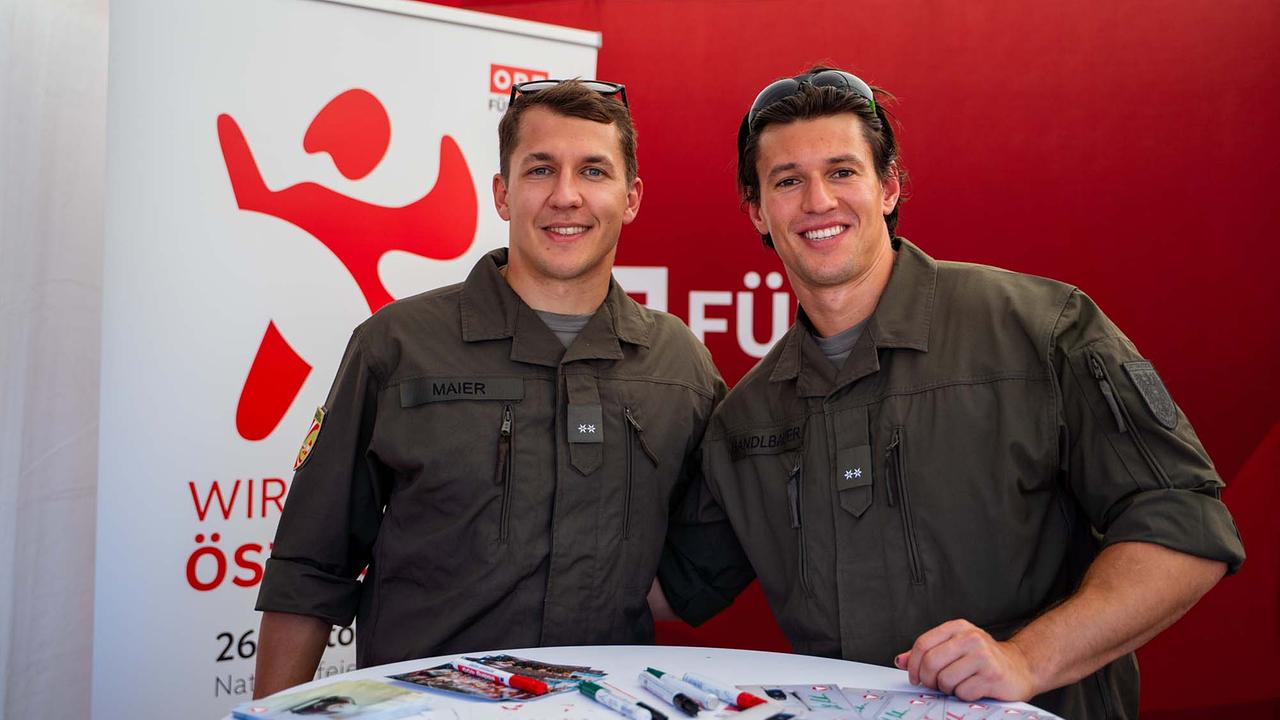 © ORF/Osman Cetin Zwei junge Männer in grünen Bundesheer-Uniform posieren im ORF‑Eventzelt Schulter an Schulter, die Ärmel sind hochgekrempelt, Sonnenbrillen sitzen auf dem Kopf. Vor ihnen liegen Autogrammkarten auf einem Tisch.