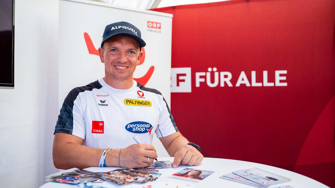 © ORF/Osman Cetin In einem ORF-Zelt steht ein Mann an einem Tisch und unterschreibt Autogrammkarten. Er trägt ein weißes Sportshirt mit Logos.
