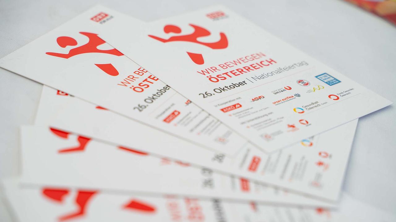 © ORF/Osman Cetin Mehrere weiße Info‑Flyer liegen fächerförmig auf einem Tisch. Im Mittelpunkt steht der rote Titel „WIR BEWEGEN ÖSTERREICH“ mit Terminangabe „26. Oktober | Nationalfeiertag“, dazu das dynamische rote Figurenlogo und ORF‑Branding. Im unteren Bereich sind Partnerlogos und Hinweise abgedruckt, unter anderem von ASKÖ, ASVÖ, SPORTUNION, Sport Austria und weiteren Institutionen.