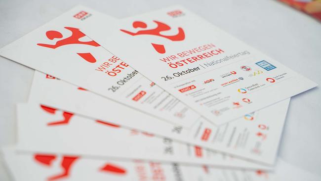 Mehrere weiße Info‑Flyer liegen fächerförmig auf einem Tisch. Im Mittelpunkt steht der rote Titel „WIR BEWEGEN ÖSTERREICH“ mit Terminangabe „26. Oktober | Nationalfeiertag“, dazu das dynamische rote Figurenlogo und ORF‑Branding. Im unteren Bereich sind Partnerlogos und Hinweise abgedruckt, unter anderem von ASKÖ, ASVÖ, SPORTUNION, Sport Austria und weiteren Institutionen.