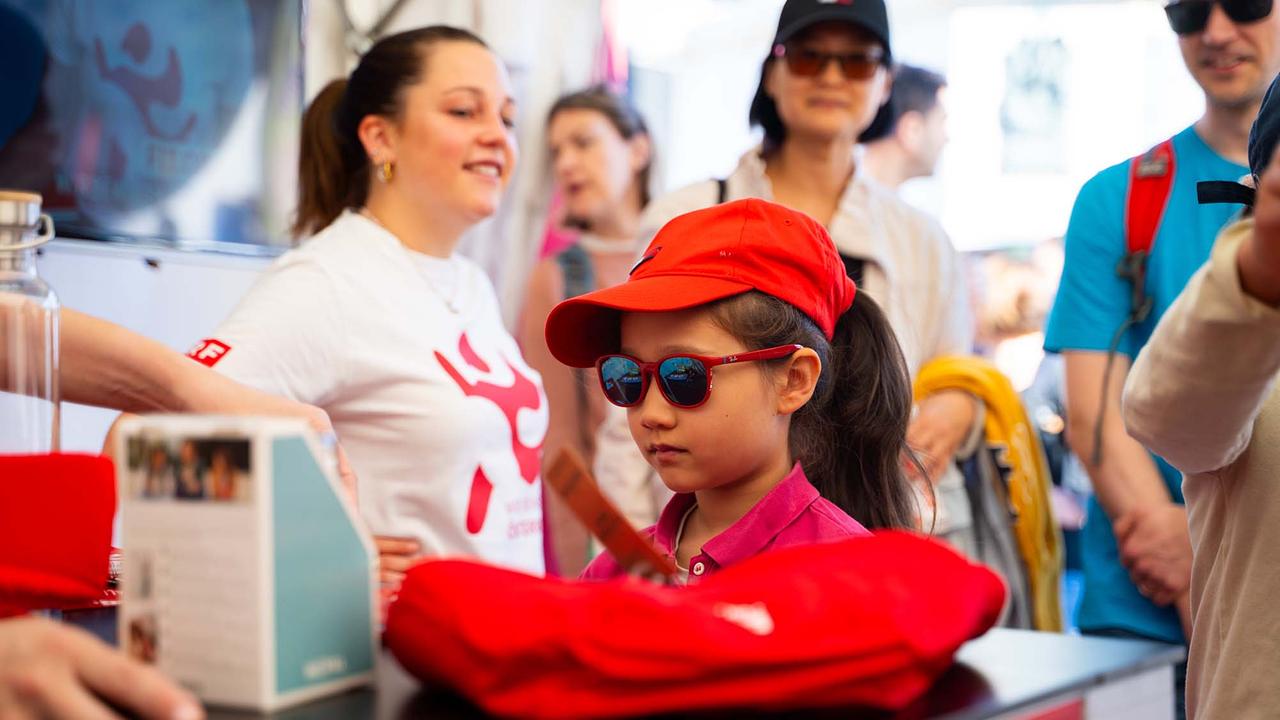 © ORF/Osman Cetin Das Bild zeigt ein Mädchen mit einer roten Jacke und einer roten Kappe. Das Mädchen steht vor einem Tisch mit Artikeln und sucht sich gerade einen aus. Im Hintergrund sieht man Menschen, die beim Glücksrad warten.