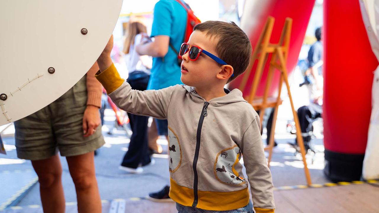 © ORF/Osman Cetin Das Bild zeigt ein Kind, das gerade ein großes Glücksrad anfasst. Der Junge trägt eine beige Kapuzenjacke mit gelben Bündchen und roten sowie blauen Sonnenbrillen. Er schaut aufmerksam nach oben in Richtung des Glücksrads. Im Hintergrund sind weitere Personen, bunte Stände und Elemente der Veranstaltung zu sehen.