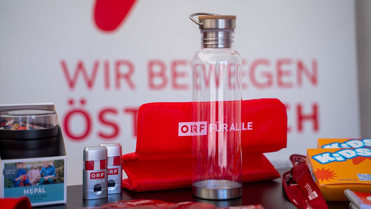 © ORF/Osman Cetin Das Bild zeigt einen Tisch mit verschiedenen Werbeartikeln des ORF. Im Mittelpunkt steht eine durchsichtige Trinkflasche mit Holzdeckel und dem Schriftzug „ORF FÜR ALLE“. Dahinter liegt ein zusammengefaltetes rotes Sitzkissen mit demselben Logo. Daneben sind kleine Behälter, Schlüsselbänder, bunte Broschüren mit der Aufschrift „KIDS“ sowie weitere Giveaways zu sehen. Im Hintergrund ist unscharf der Schriftzug „WIR BEWEGEN ÖSTERREICH“ erkennbar.