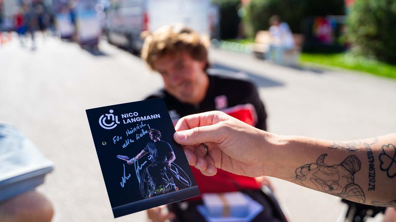 © ORF/Osman Cetin Das Bild zeigt eine Nahaufnahme einer Hand, die eine signierte Autogrammkarte von Nico Langmann hält. Darauf ist der Rollstuhltennisspieler beim Schlag abgebildet, daneben steht eine persönliche Widmung. Im Hintergrund, leicht unscharf, sitzt Nico Langmann selbst im Rollstuhl und lächelt. Im Vordergrund ist zudem der Arm der Person zu sehen, die die Karte hält, mit einem auffälligen Tattoo.