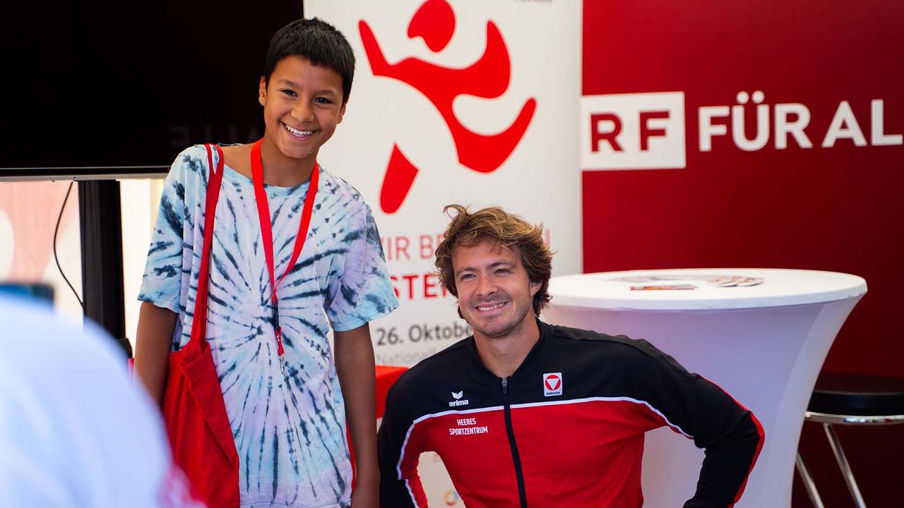 © ORF/Osman Cetin Das Bild zeigt Nico Langmann (Profi-Tennisspieler) und einen jungen Besucher am Tag des Sports in der ORF-Pagode. Im Hintergrund sieht man ein Werbeschild ORF FÜR ALLE. Nico Langmann sitzt im Rollstuhl mit einem rot-schwarzen Trainingsanzug mit der Aufschrift Austria. Der junge Besucher steht rechts von Nico und hat ein hellblaues T-Shirt an und ein rotes Lanyard und eine rote Tasche über der rechten Schulter. Beide Personen strahlen.
