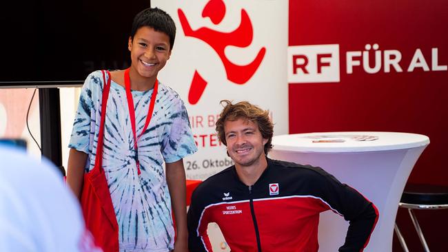 Das Bild zeigt Nico Langmann (Profi-Tennisspieler) und einen jungen Besucher am Tag des Sports in der ORF-Pagode. Im Hintergrund sieht man ein Werbeschild ORF FÜR ALLE. Nico Langmann sitzt im Rollstuhl mit einem rot-schwarzen Trainingsanzug mit der Aufschrift Austria. Der junge Besucher steht rechts von Nico und hat ein hellblaues T-Shirt an und ein rotes Lanyard und eine rote Tasche über der rechten Schulter. Beide Personen strahlen.
