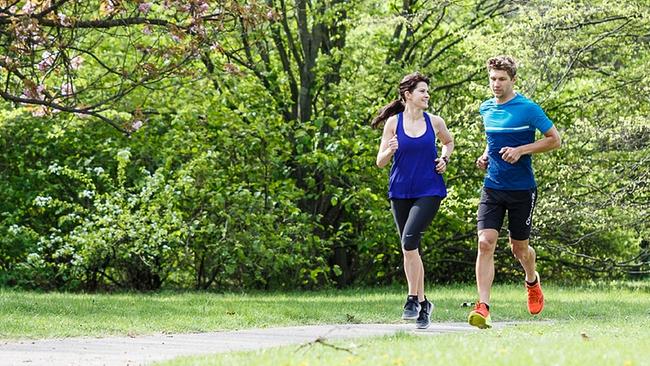 Eine Joggerin und ein Jogger laufen nebeneinander auf einem Weg, um sie herum ist es grün, man sieht Bäume und Sträucher.